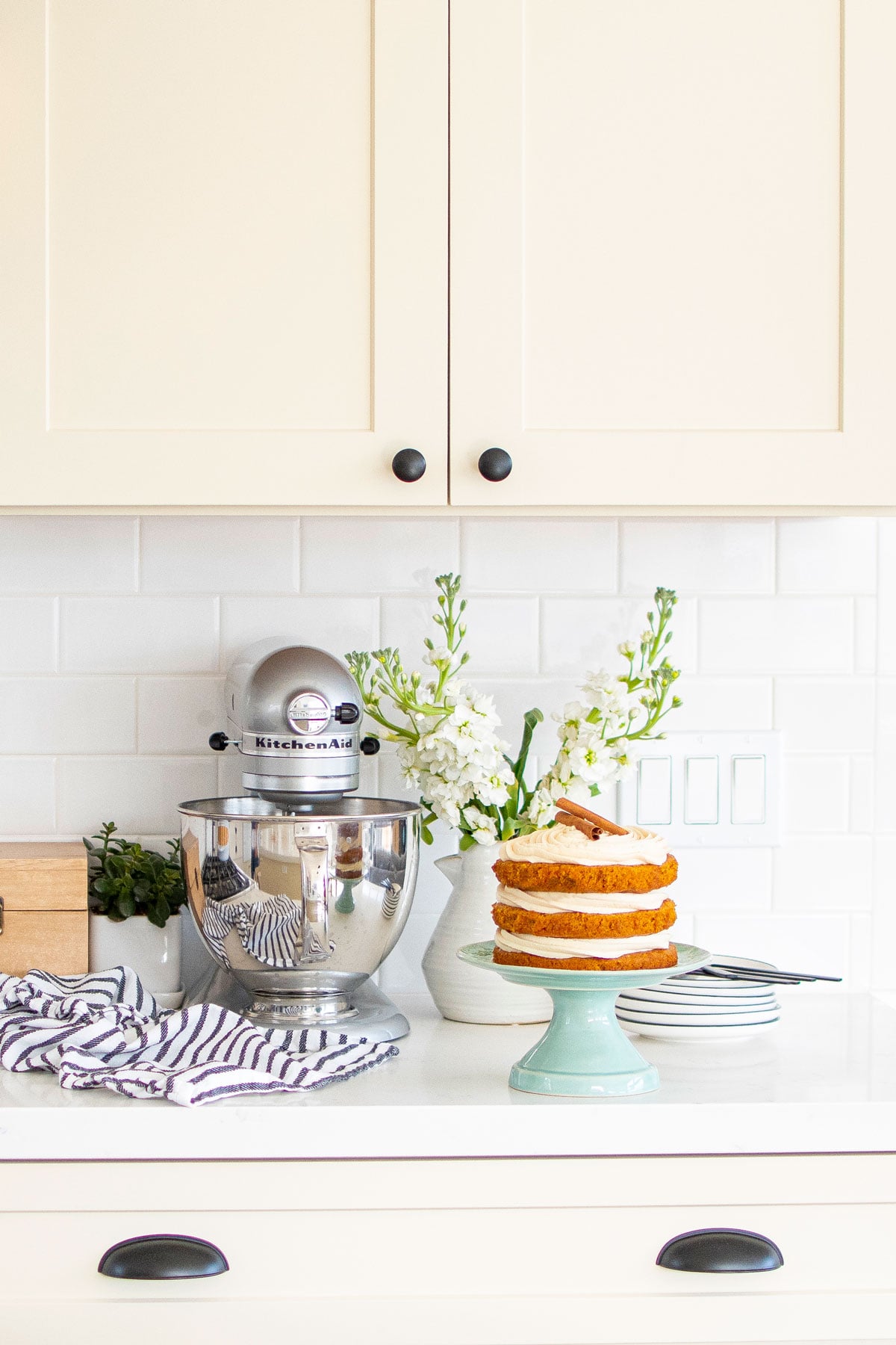 silver stand mixer on a white kitchen counter with a naked carrot cake on a green cake stand.