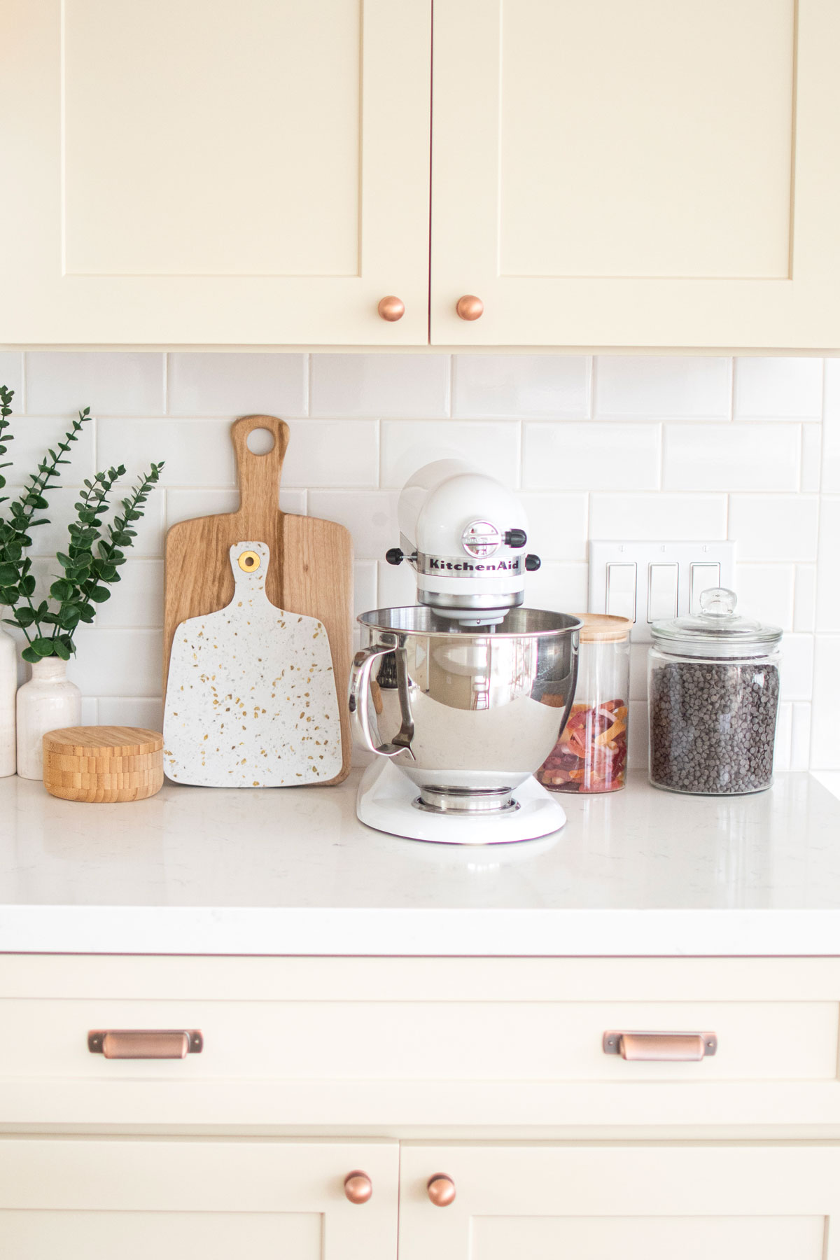 white kitchenaid stand mixer on a white kitchen counter with cutting boards and glass jars of sweets.