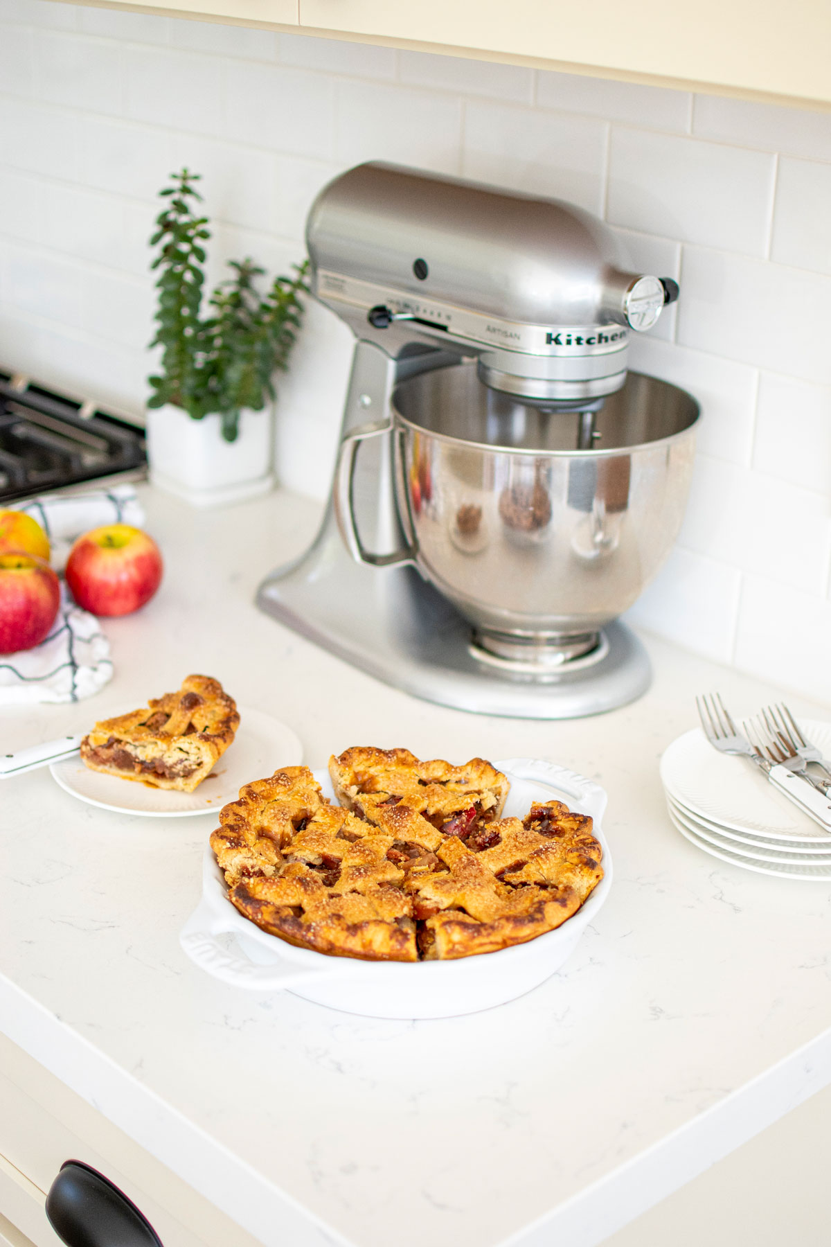 lattice apple pie cut into slices on a white kitchen counter with a stainless steel stand mixer.