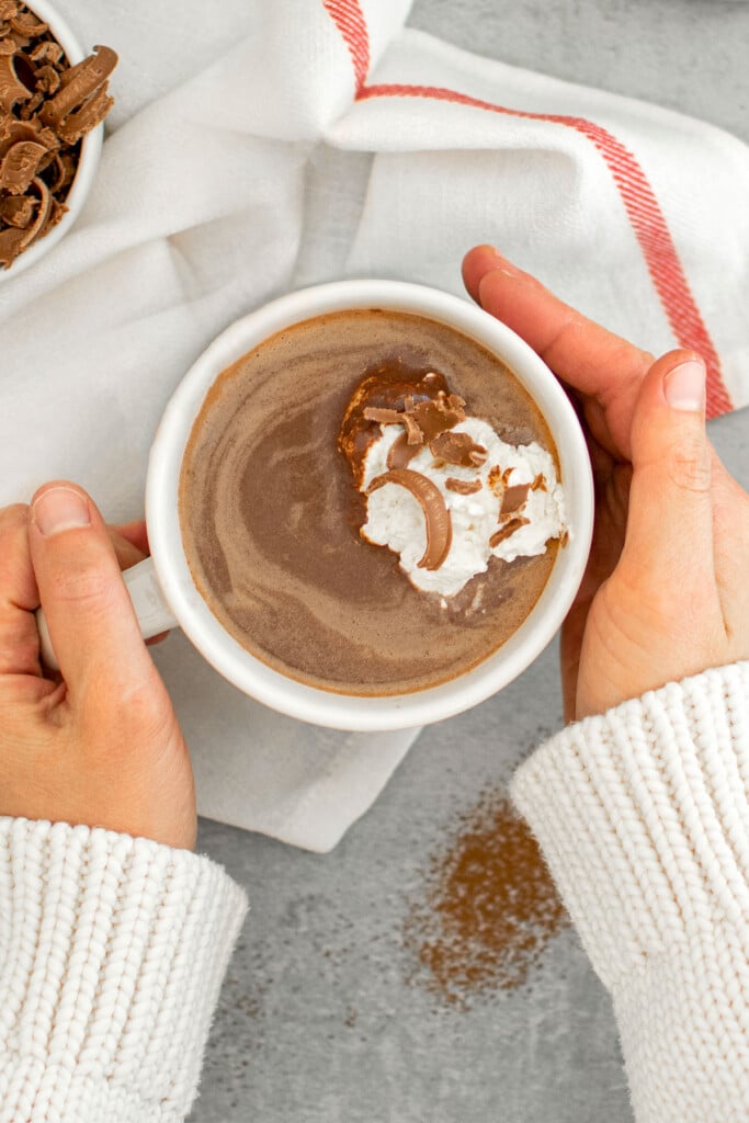 woman holding a mug of dark hot chocolate with whipped cream and chocolate shavings on top.