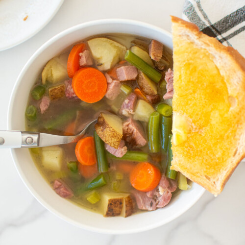 ham bone soup in a bowl with a slice of toast on the side on a white marble countertop.