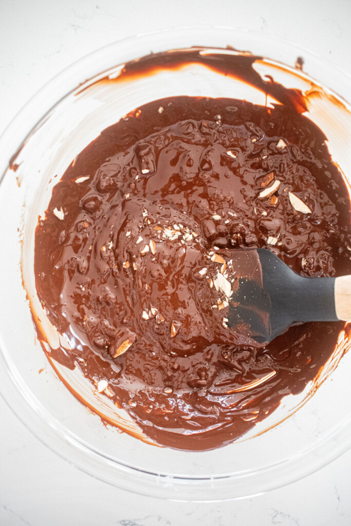 melted chocolate with almonds in a glass mixing bowl on a white marble counter.