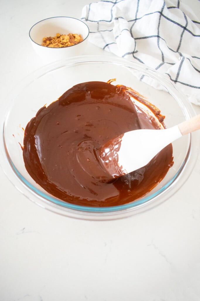 melted dark chocolate in a glass mixing bowl with a spatula on a marble counter with a small bowl of chopped almonds.