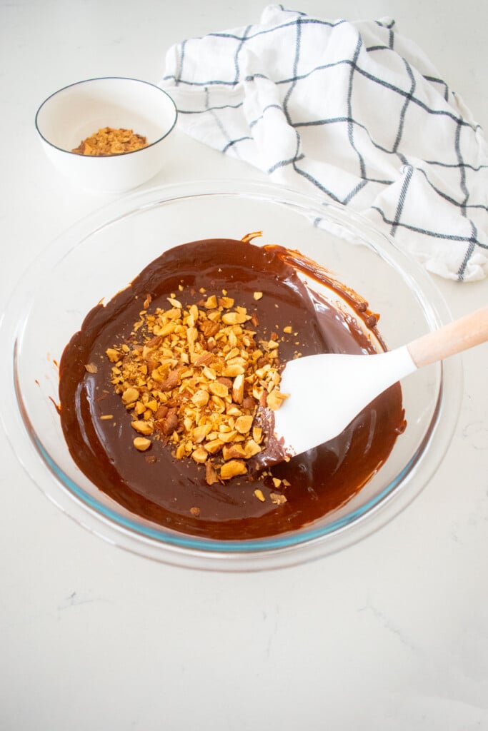 glass mixing bowl with melted dark chocolate and chopped almonds with a spatula on a marble counter.
