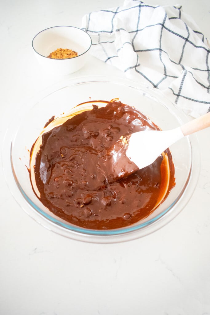 melted dark chocolate with almonds mixed into it in a glass mixing bowl with a spatula on a marble counter.