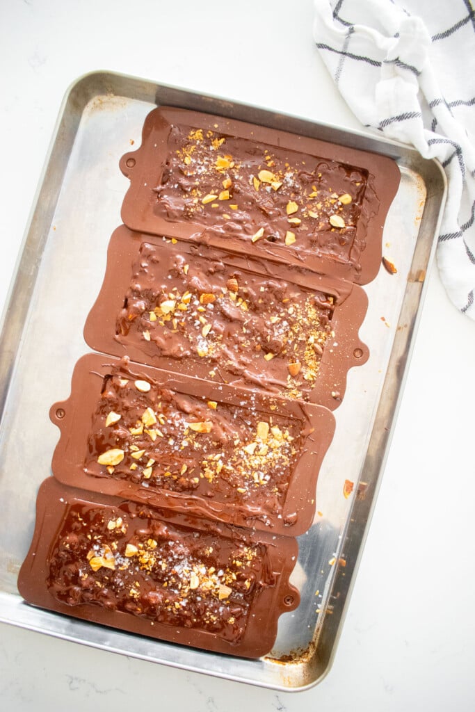 chocolate bar molds filled with dark chocolate and almonds on a baking sheet on a marble counter.
