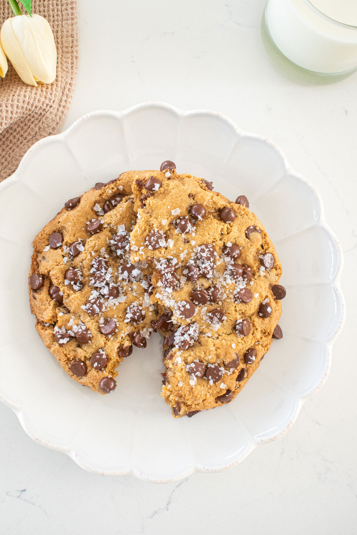 single serve chocolate chip cookie with sea salt broke in half on a plate on a marble counter with a glass of milk and fresh flowers.
