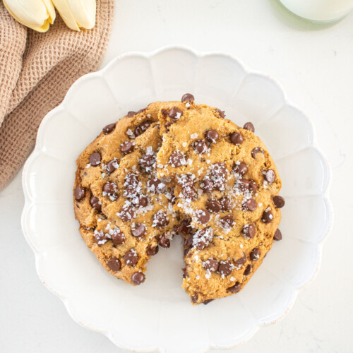 single serve chocolate chip cookie with sea salt broke in half on a plate on a marble counter with a glass of milk and fresh flowers.