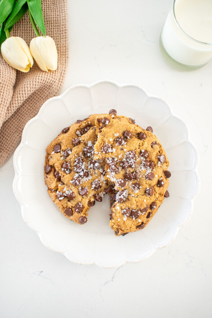 single serve chocolate chip cookie with sea salt broke in half on a plate on a marble counter with a glass of milk and fresh flowers.
