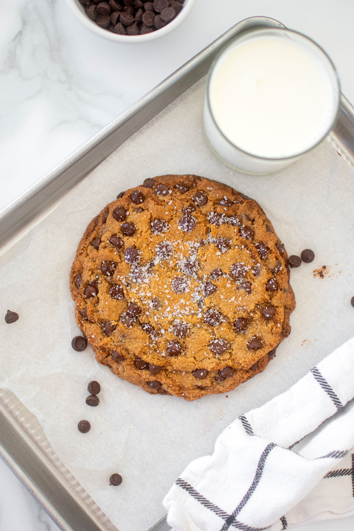 single serve chocolate chip cookie with flaky sea salt on a small baking sheet with a glass of milk on a marble counter.