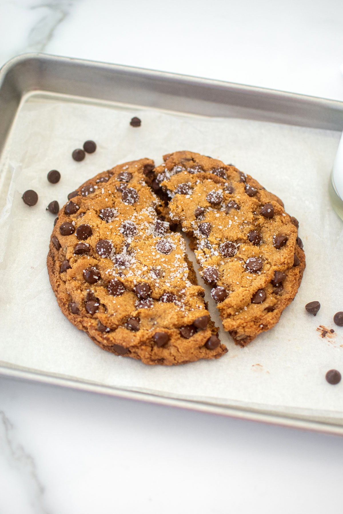 single serve chocolate chip cookie broke in half on a small baking sheet on a marble counter.