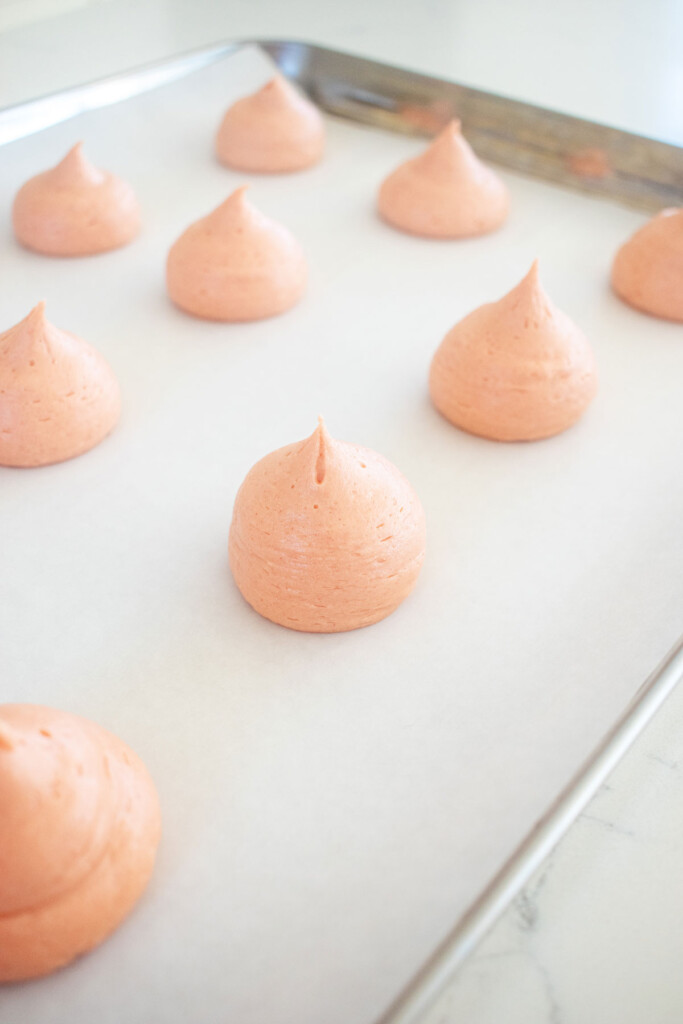 piped dollops of strawberry cream drops on a baking sheet on a white marble counter.
