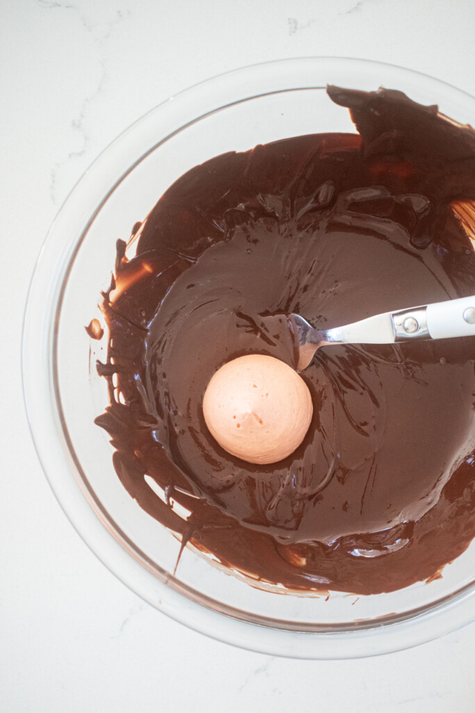 strawberry cream drop being coated in dark chocolate in a glass mixing bowl on a white marble counter.