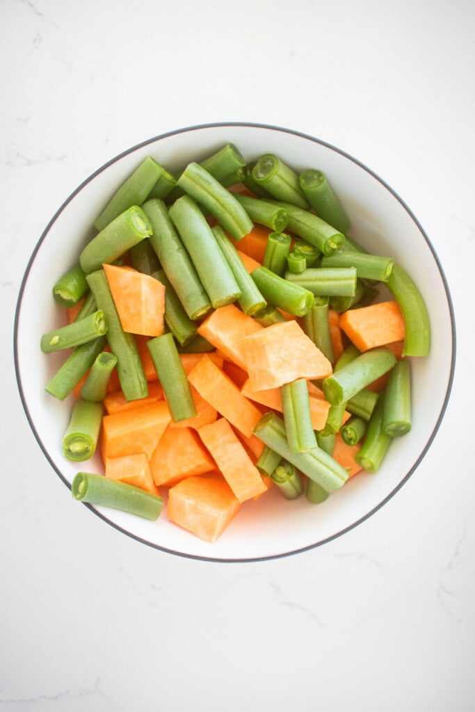 peeled sweet potatoes and chopped green beans in a bowl on a white countertop.