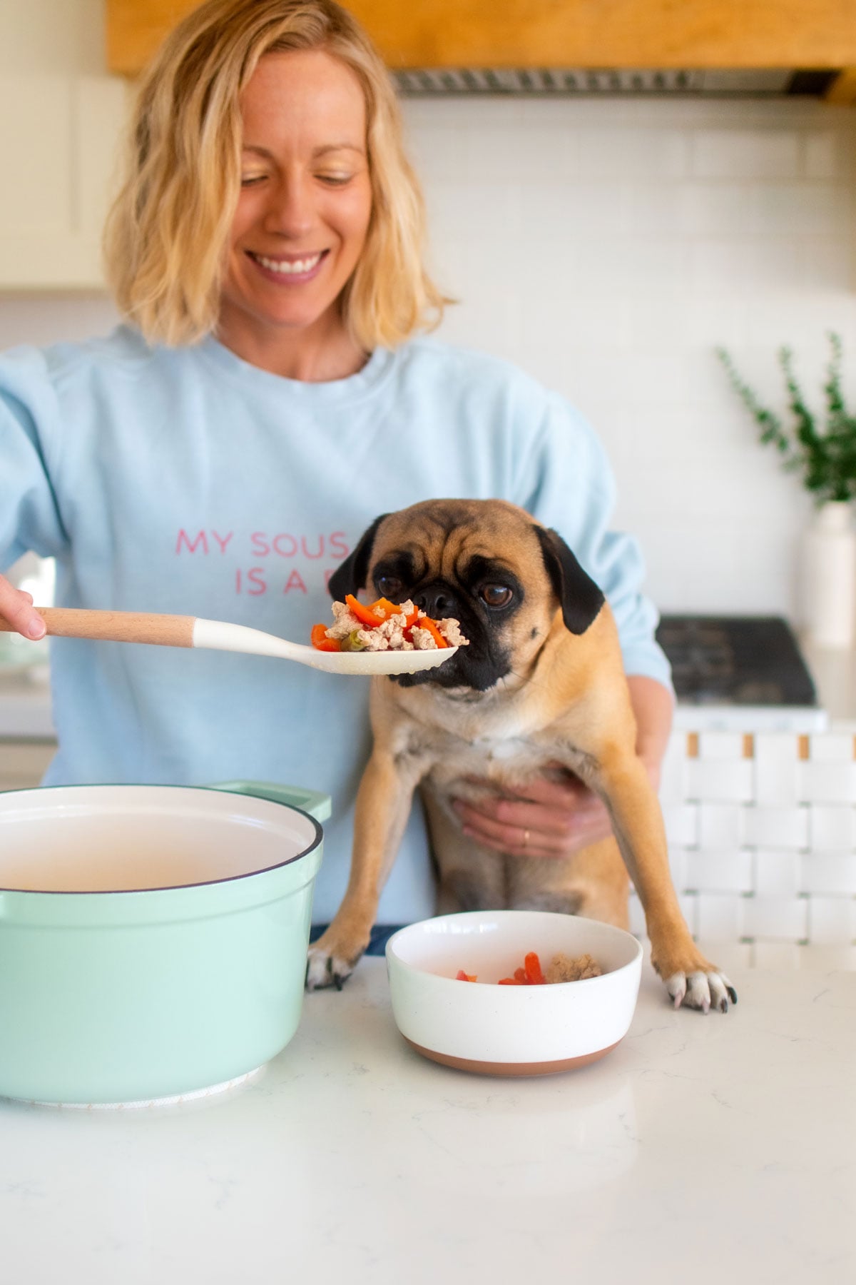 woman serving homemade turkey dog food to a pug at the kitchen counter.