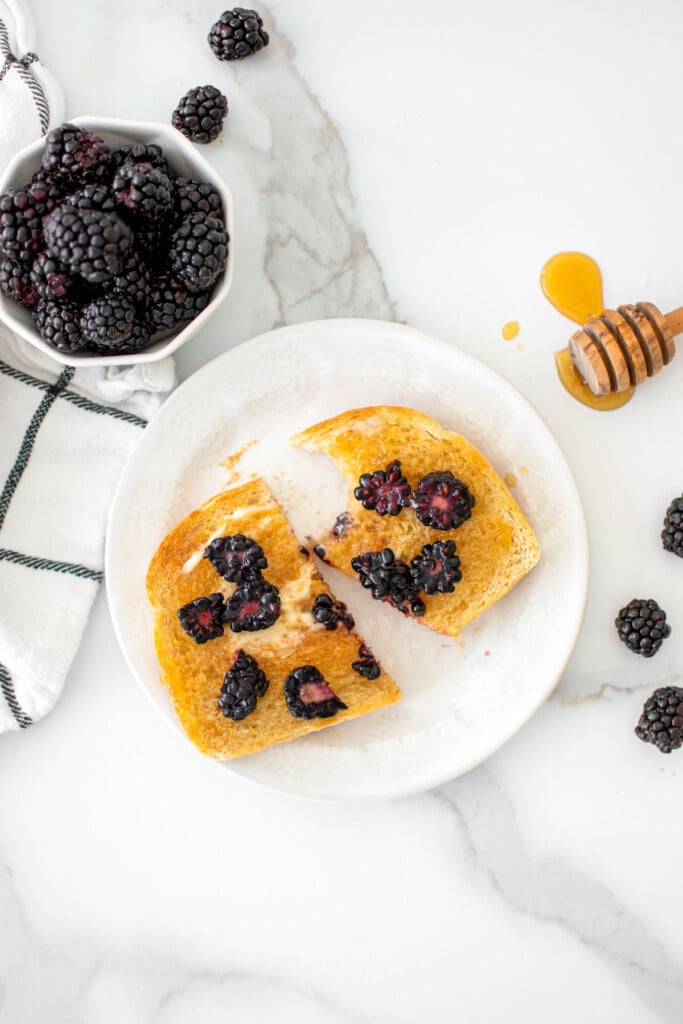 blackberry honey butter toast cut in half on a white plate on a marble countertop with a bowl of blackberries.