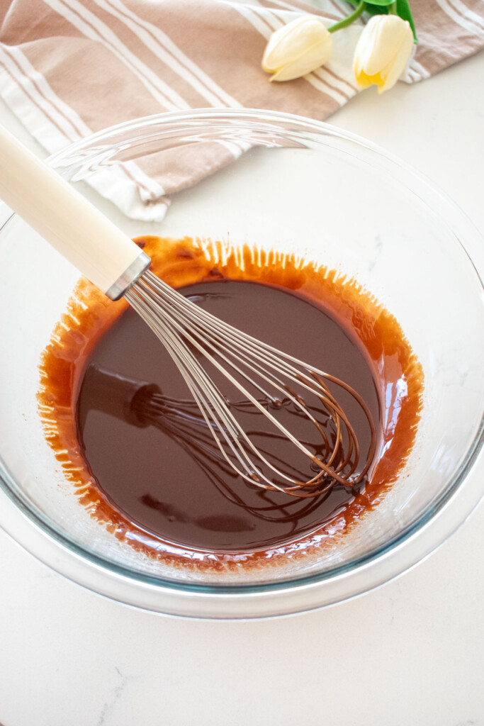melted butter and chocolate in a glass mixing bowl with a whisk on a white marble counter.