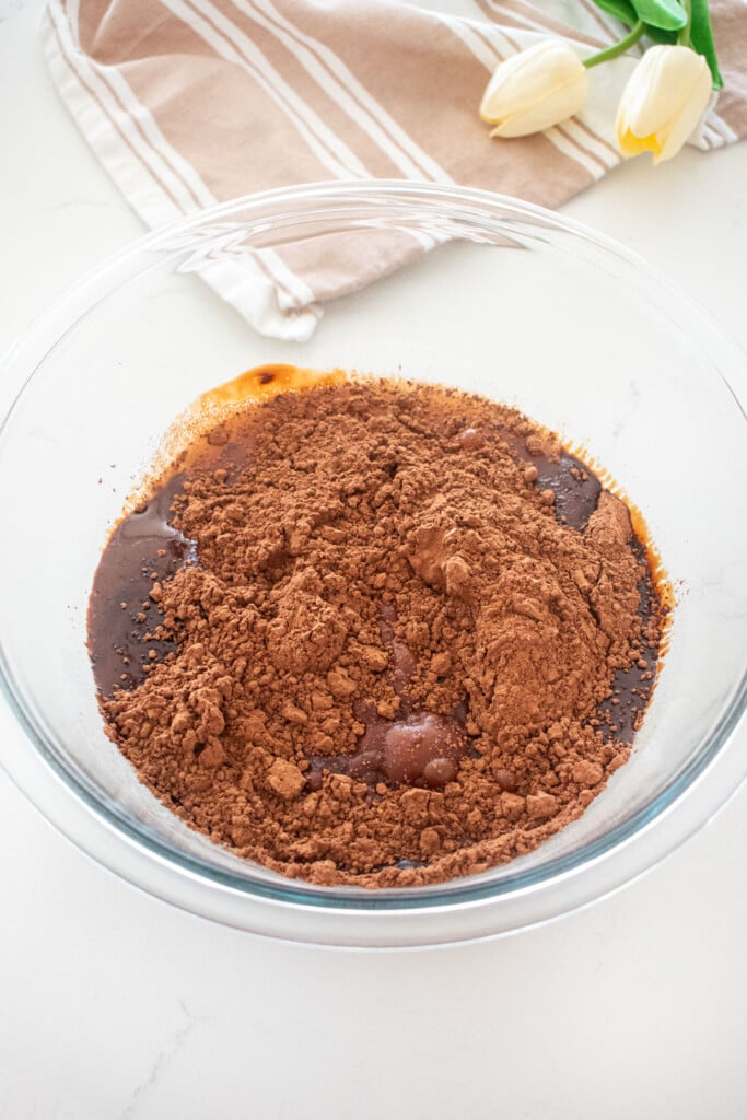brownie batter ingredients being added to a glass mixing bowl on a white marble counter.