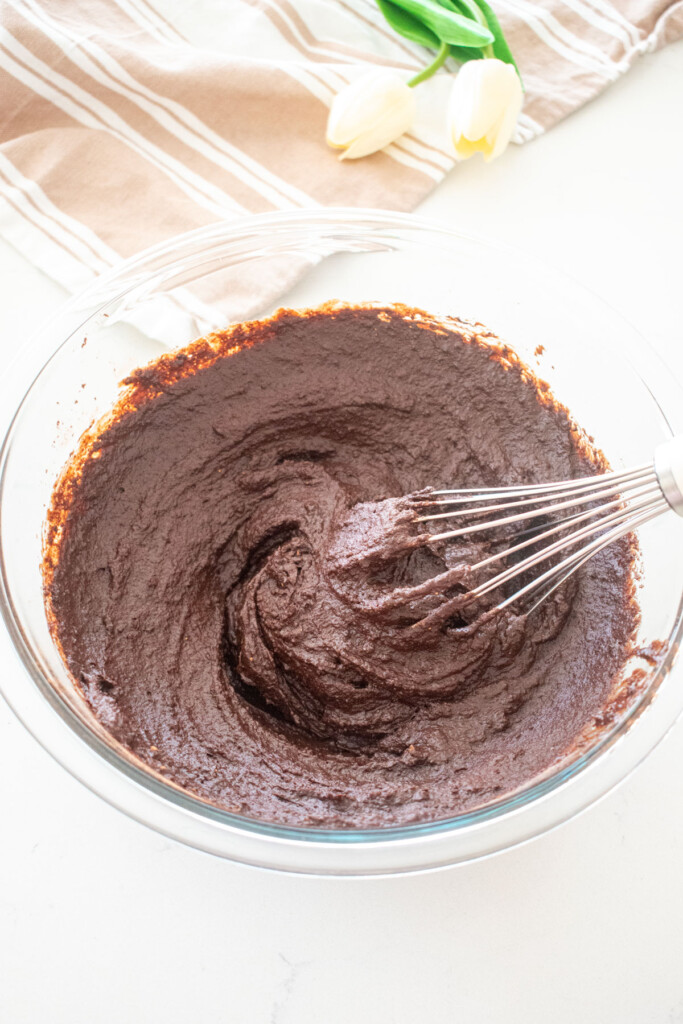 brownie batter being whisked together in a glass mixing bowl on a white marble counter.