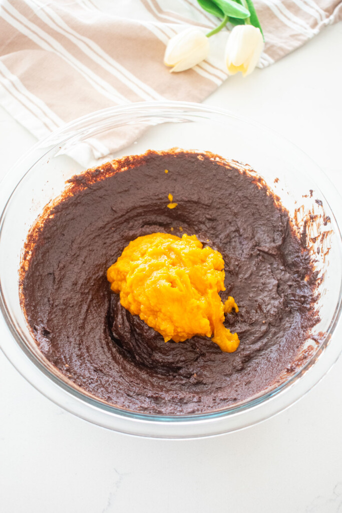 brownie batter with pumpkin puree being added to it in a glass mixing bowl on a white marble counter.