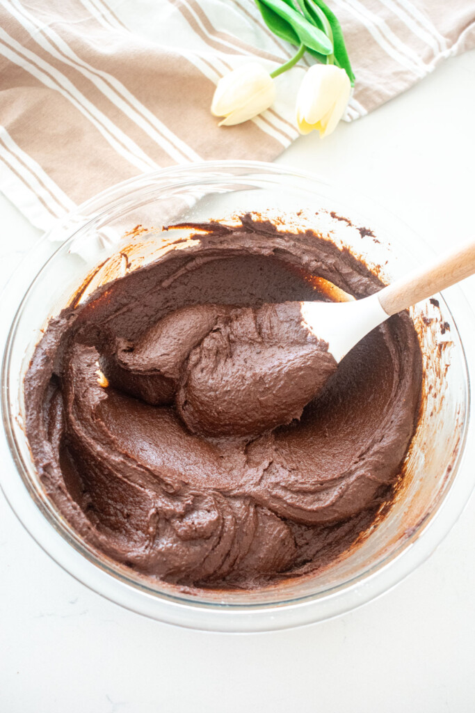 smooth brownie batter being stirred with a spatula in a glass mixing bowl on a white marble counter.