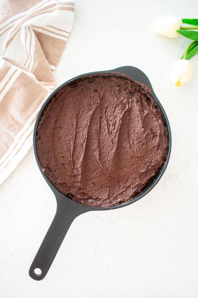brownie batter in a cast iron skillet on a white marble counter with fresh flowers.