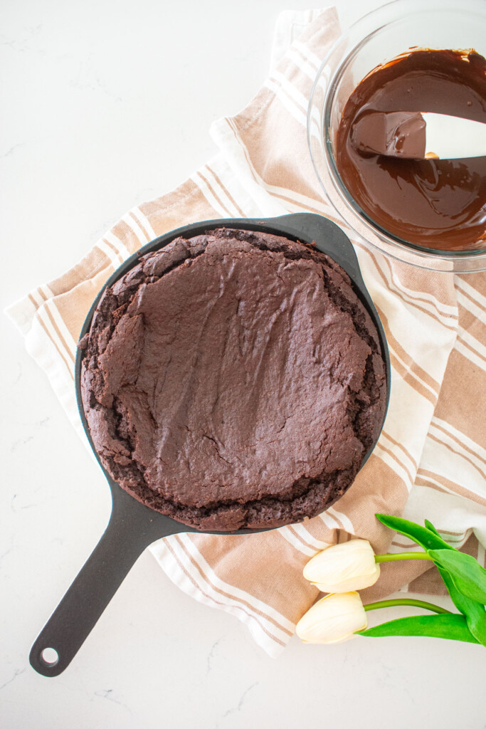 baked brownie skillet on a white marble counter with a bowl of melted chocolate next it and white tulips.