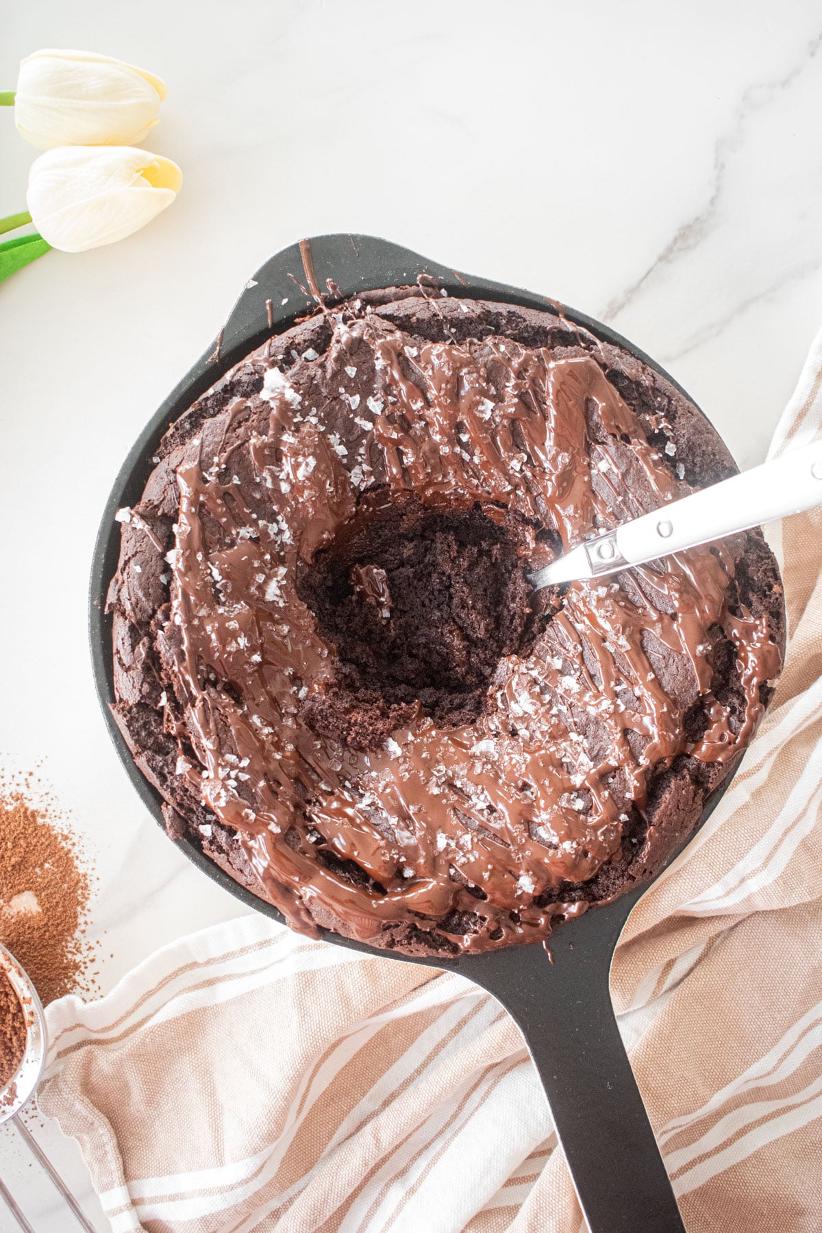 bone broth brownies in a skillet with melted chocolate drizzle on top and flakey sea salt on a white marble counter with a spoon in the middle with a bite taken out.