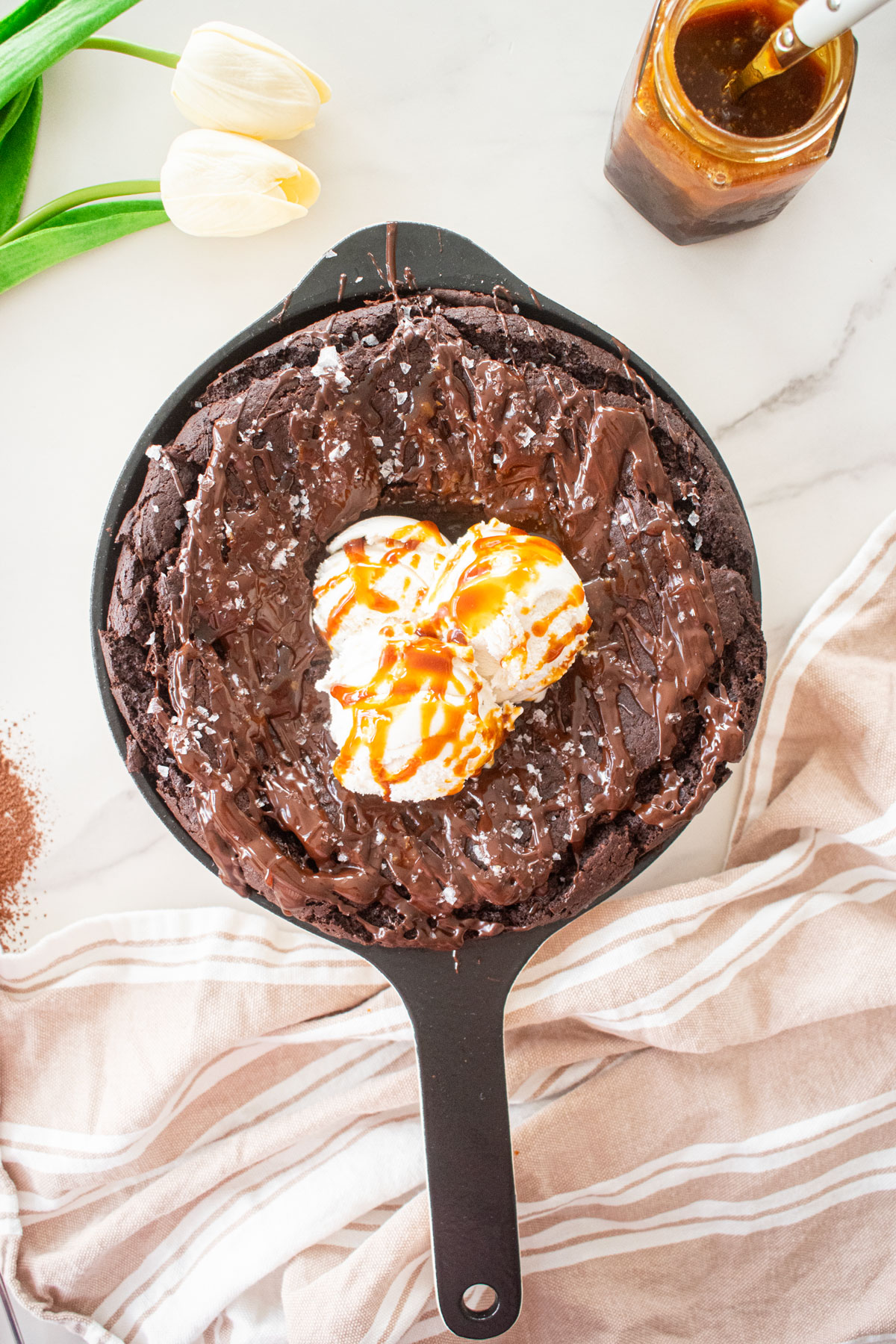 bone broth brownies skillet with melted chocolate, flaky sea salt and 3 scoops of vanilla ice cream with caramel drizzle on a white marble counter.