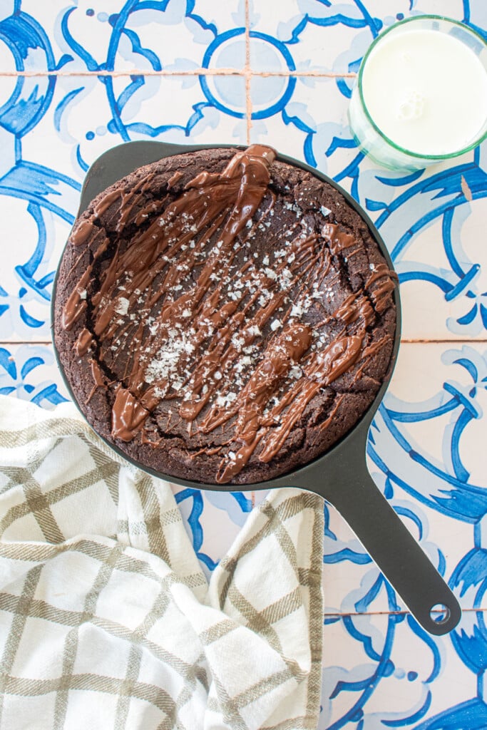 bone broth brownies in a skillet with melted chocolate and flaky sea salt on a blue tile table with a glass of milk.