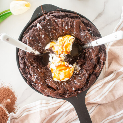 bone broth brownies skillet with melted chocolate, flaky sea salt and 3 scoops of vanilla ice cream with caramel drizzle and two spoons on a white marble counter.