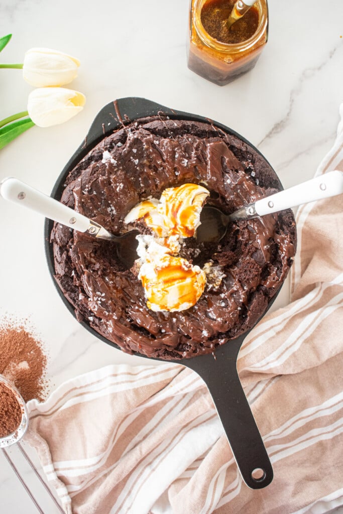 bone broth brownies skillet with melted chocolate, flaky sea salt and 3 scoops of vanilla ice cream with caramel drizzle and two spoons on a white marble counter.