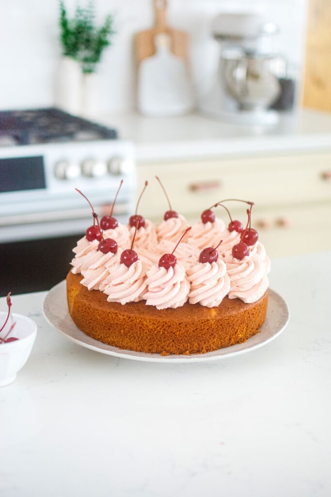 cherry cake with whipped cream frosting and marschino cherries on top on a cake plate on the counter.