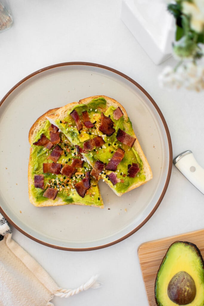 everything bacon avocado toast on sourdough bread cut in half on a gray plate on marble countertop.