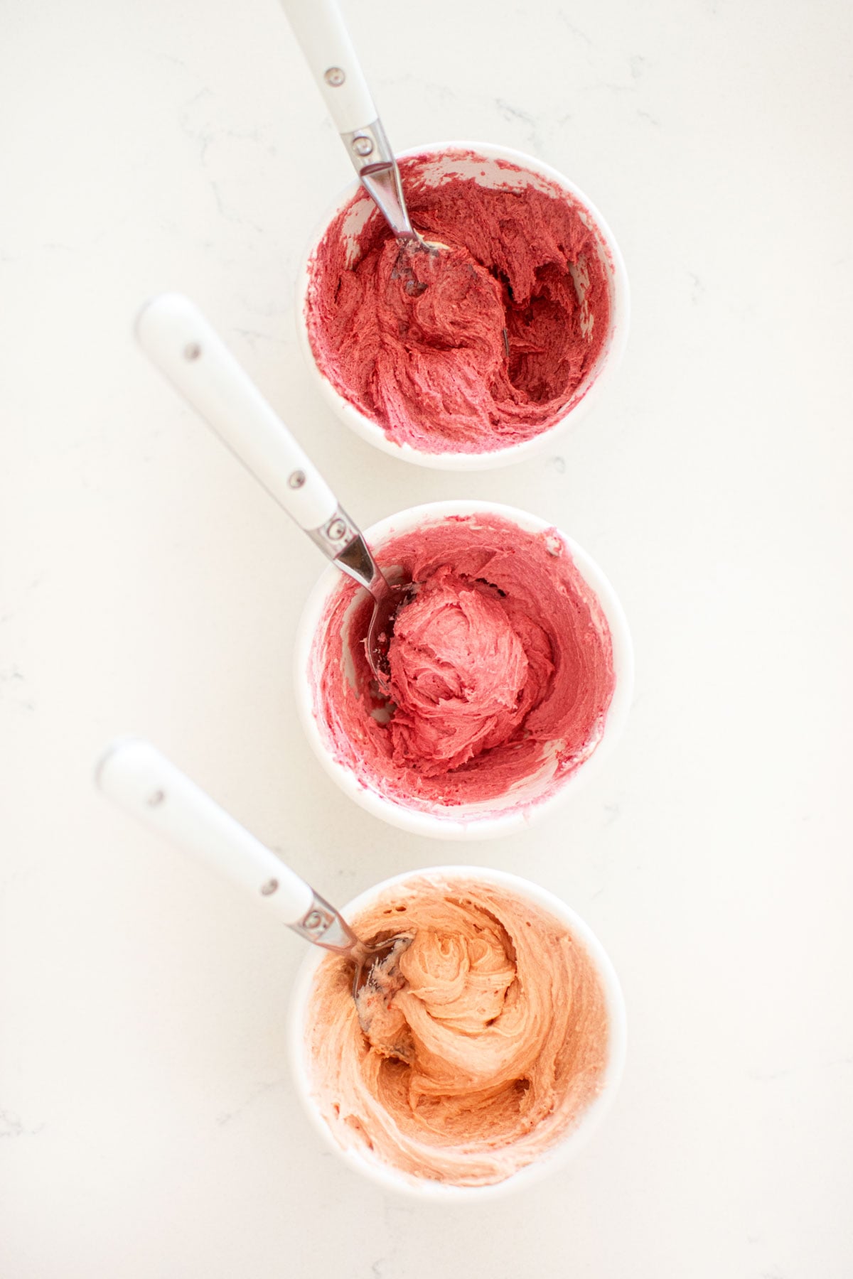 three small bowls of different shades of pink frosting on a white marble counter.