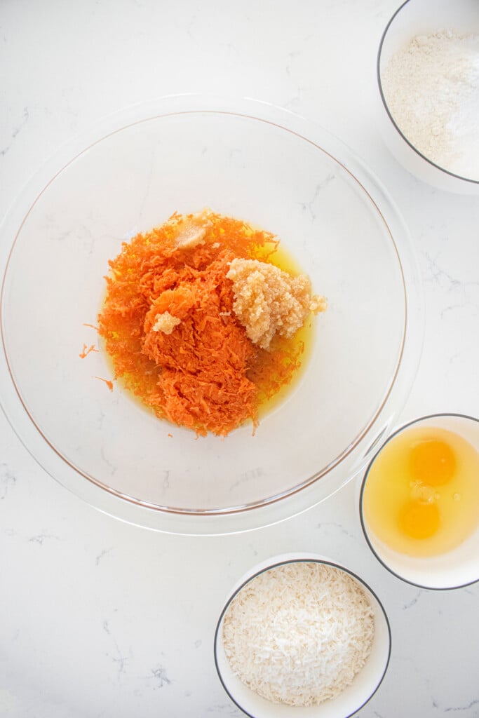 carrots, oil, and honey in a glass mixing bowl on a white countertop.