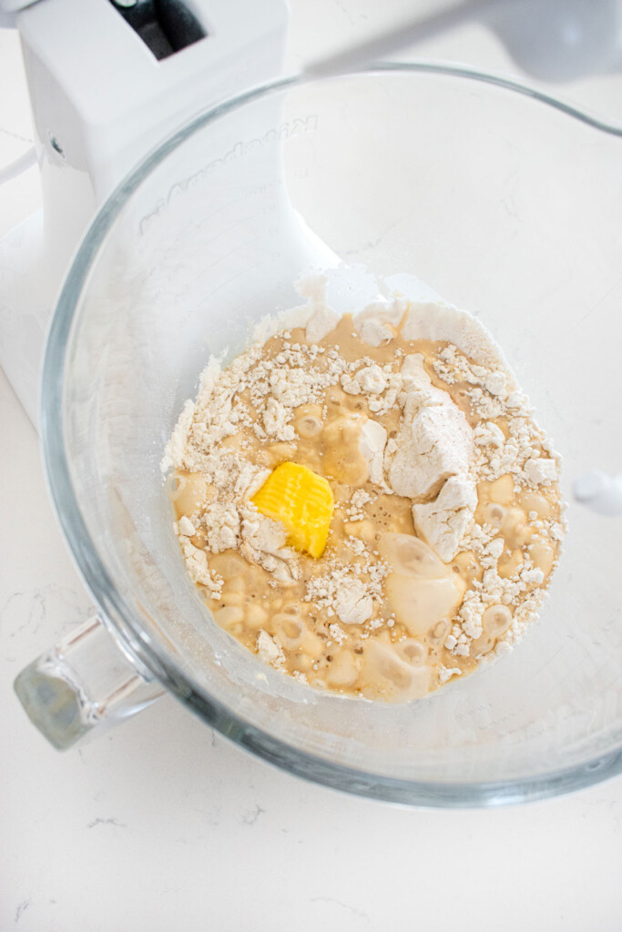 ingredients for sourdough hot dog pretzel bun dough in a glass mixing bowl on a white marble countertop.