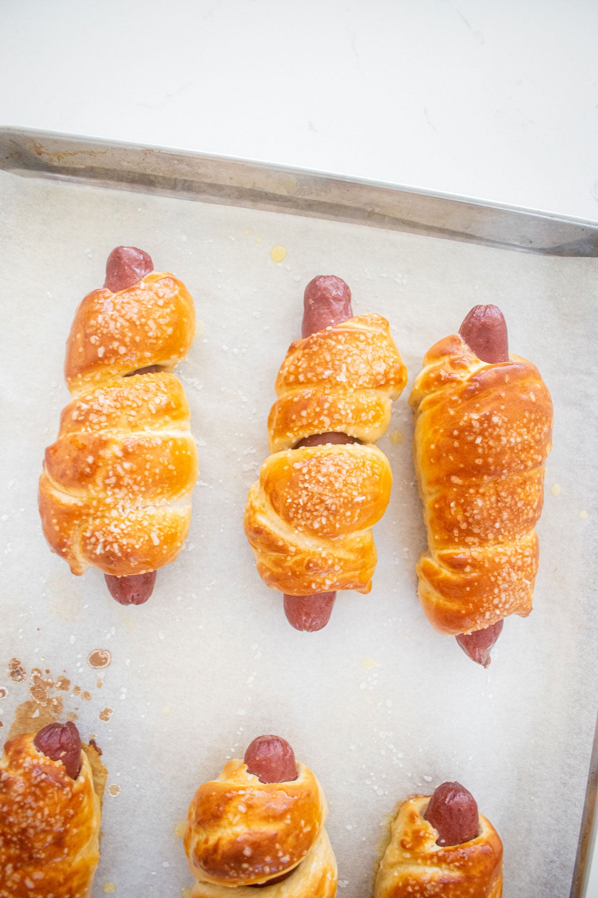 baked sourdough hot dog pretzel buns on a baking sheet with butter brushed on them on a marble countertop.
