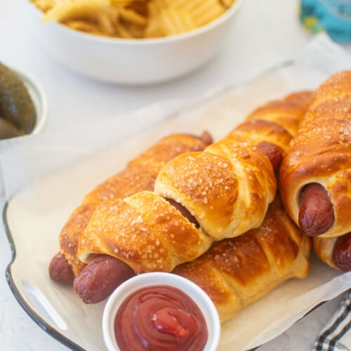 sourdough hot dog pretzel buns on a plate with a plate of chips, pickles and a beer.
