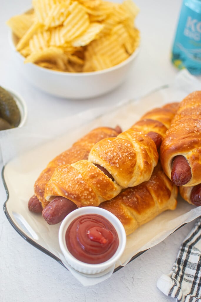 sourdough hot dog pretzel buns on a plate with a plate of chips, pickles and a beer.