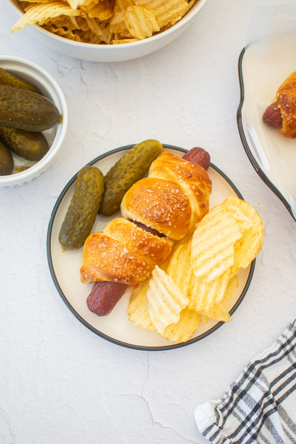 sourdough hot dog pretzel bun on a plate with pickles and chips.