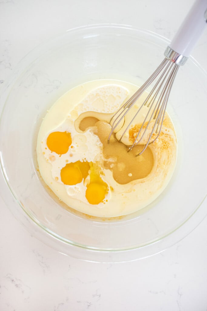 wet ingredients for vanila cake in a glass mixing bowl being whisked together on a white marble counter.
