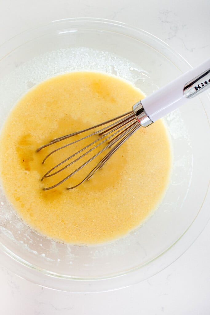 ingredients for cookie dough layer cake in a glass mixing bowl with a whisk on a white marble counter.