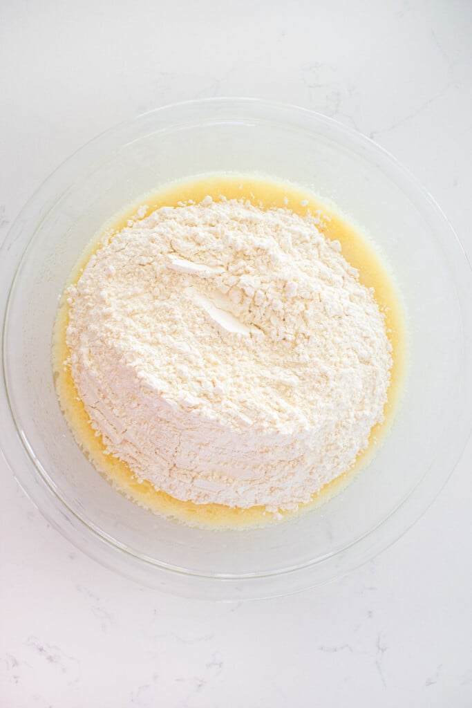 dry ingredients being added into wet ingredients for vanilla cake bater in a glass mixing bowl on a marble counter.