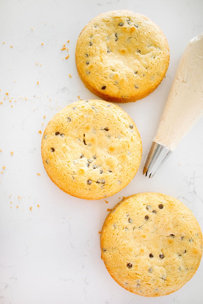 baked cookie dough cake layers on a white marble counter with cookie dough frosting in a piping bag.
