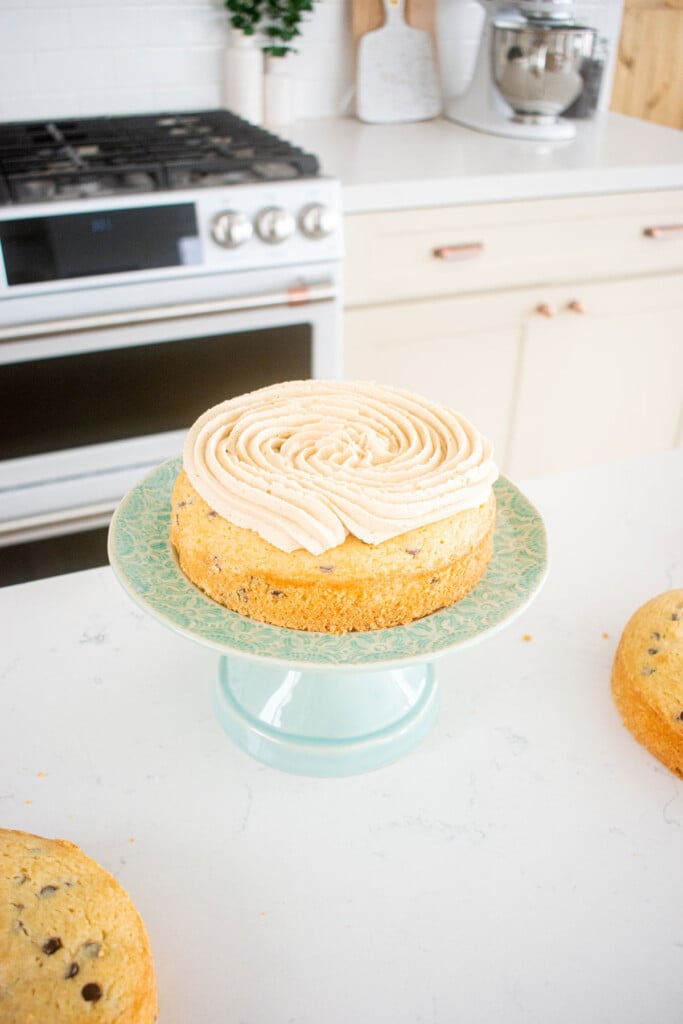 cookie dough layer cake being assembled on a green cake stand on the kitchen counter.