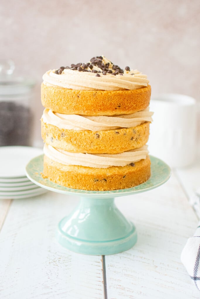 cookie dough layer cake with mini chocolate chips on top sitting on a green cake stand on a white wood table.