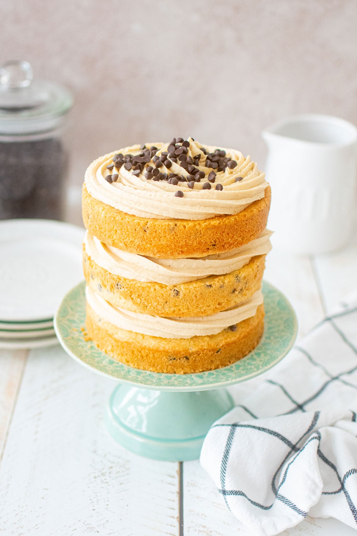 cookie dough layer cake with mini chocolate chips on top sitting on a green cake stand on a white wood table.