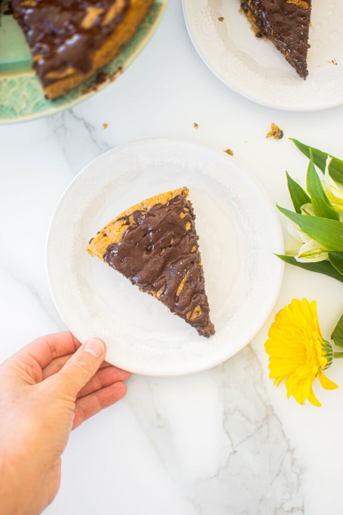 woman grabbing eggless chocolate chip cookie cake slice on a white plate on a marble counter.