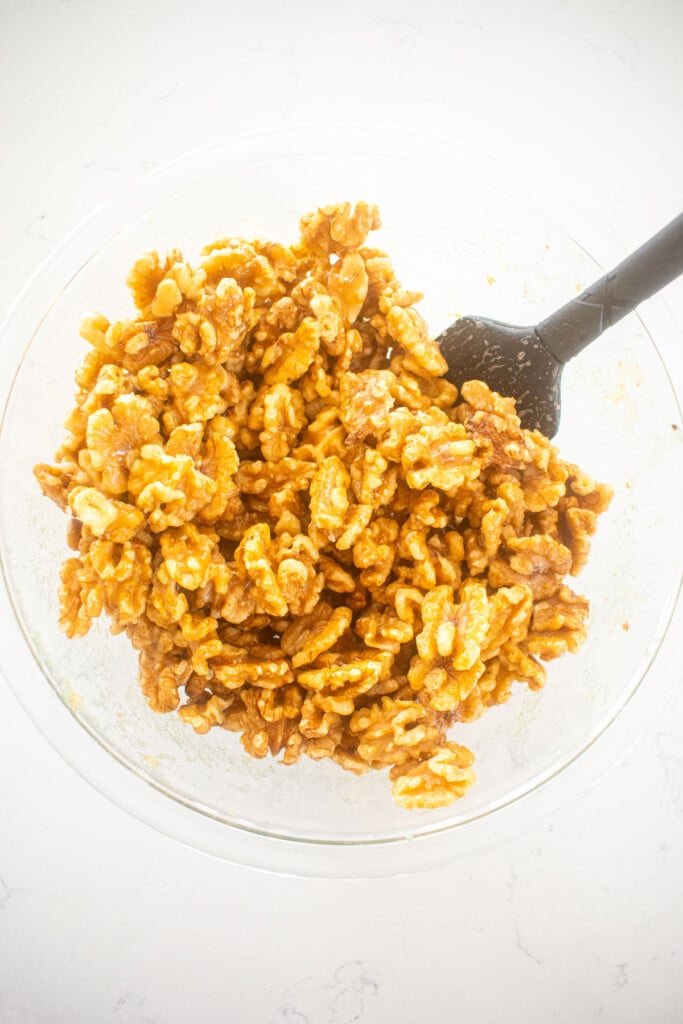 raw walnuts being mixed with hot honey in a glass bowl on a white marble countertop.
